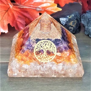 May include: A clear crystal pyramid with a gold tree of life design and layers of orange, purple, and clear crystals. The pyramid has a copper wire coil at the top.