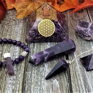 May include: A set of amethyst crystals, including a pyramid, a point, a pendulum, a bracelet, and a tumbled stone. The crystals are all a deep purple color and are arranged on a wooden surface.
