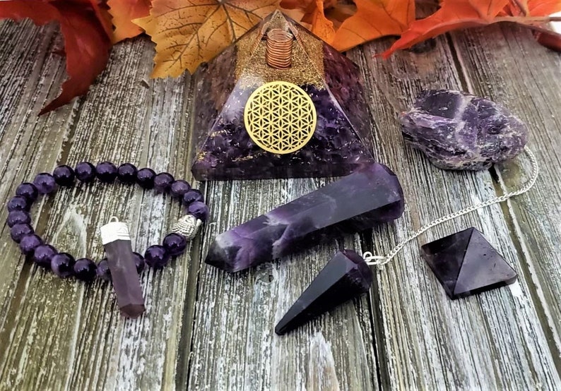 May include: A set of amethyst crystals, including a pyramid, a point, a pendulum, a bracelet, and a tumbled stone. The crystals are all a deep purple color and are arranged on a wooden surface.