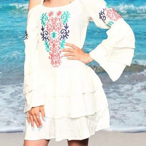 Puede incluir: Un vestido blanco de hombros descubiertos con mangas largas y falda de capas. El vestido presenta diseños florales bordados de colores en el corpiño.