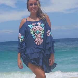 May include: A woman wearing a blue, off-the-shoulder dress with colourful embroidery. The dress has a ruffled hem and is worn over a white swimsuit. The woman is standing on a beach with the ocean in the background.