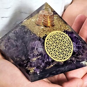 May include: Amethyst pyramid with a gold flower of life symbol and copper coils inside. The pyramid is made of clear resin and has a purple amethyst crystal base.