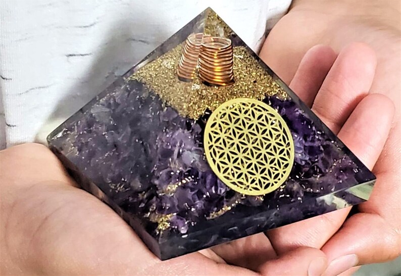 May include: Amethyst pyramid with a gold flower of life symbol and copper coils inside. The pyramid is made of clear resin and has a purple amethyst crystal base.