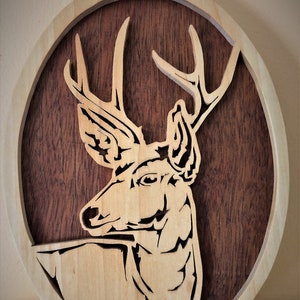 May include: A wooden wall decor featuring a cutout of a deer head with antlers. The deer is carved in a light wood and is set against a dark wood background. The piece is oval shaped and has a natural wood finish.