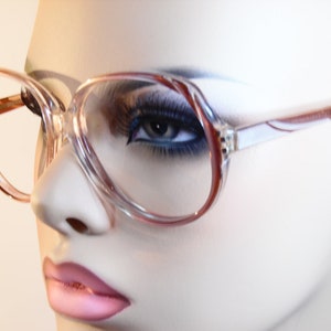 May include: Clear-framed eyeglasses with a reddish-brown accent on the upper rim and temples. The glasses have a large, rounded shape. The image shows the glasses on a mannequin head.