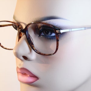 Puede incluir: Primer plano de gafas con montura de carey y varillas plateadas. Las gafas, de forma cuadrada, se presentan sobre un fondo neutro. La imagen se centra en las gafas.