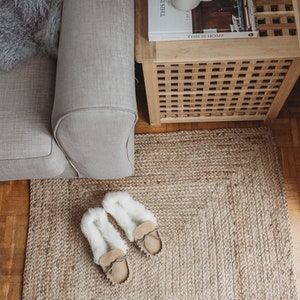 May include: A close-up of a jute rug with two pairs of fuzzy slippers on top. The rug is beige and the slippers are white with brown leather soles. The rug is in front of a couch and a wooden side table with a book titled "This is Home" on top.
