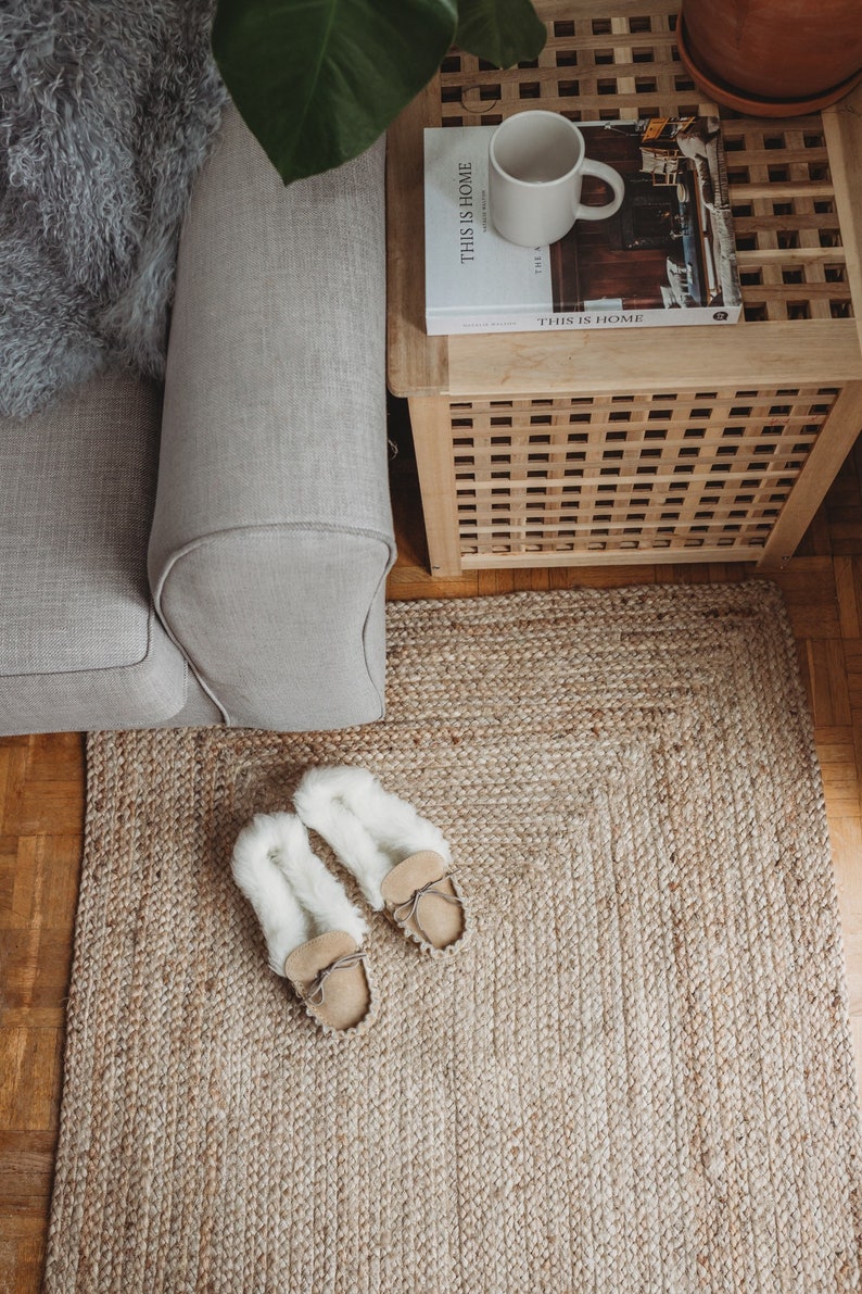 May include: A close-up of a jute rug with two pairs of fuzzy slippers on top. The rug is beige and the slippers are white with brown leather soles. The rug is in front of a couch and a wooden side table with a book titled "This is Home" on top.
