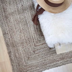 May include: A close-up of a woven jute rug with a light brown color. The rug has a textured surface and a geometric pattern. A white faux fur throw and a straw hat are on top of the rug.