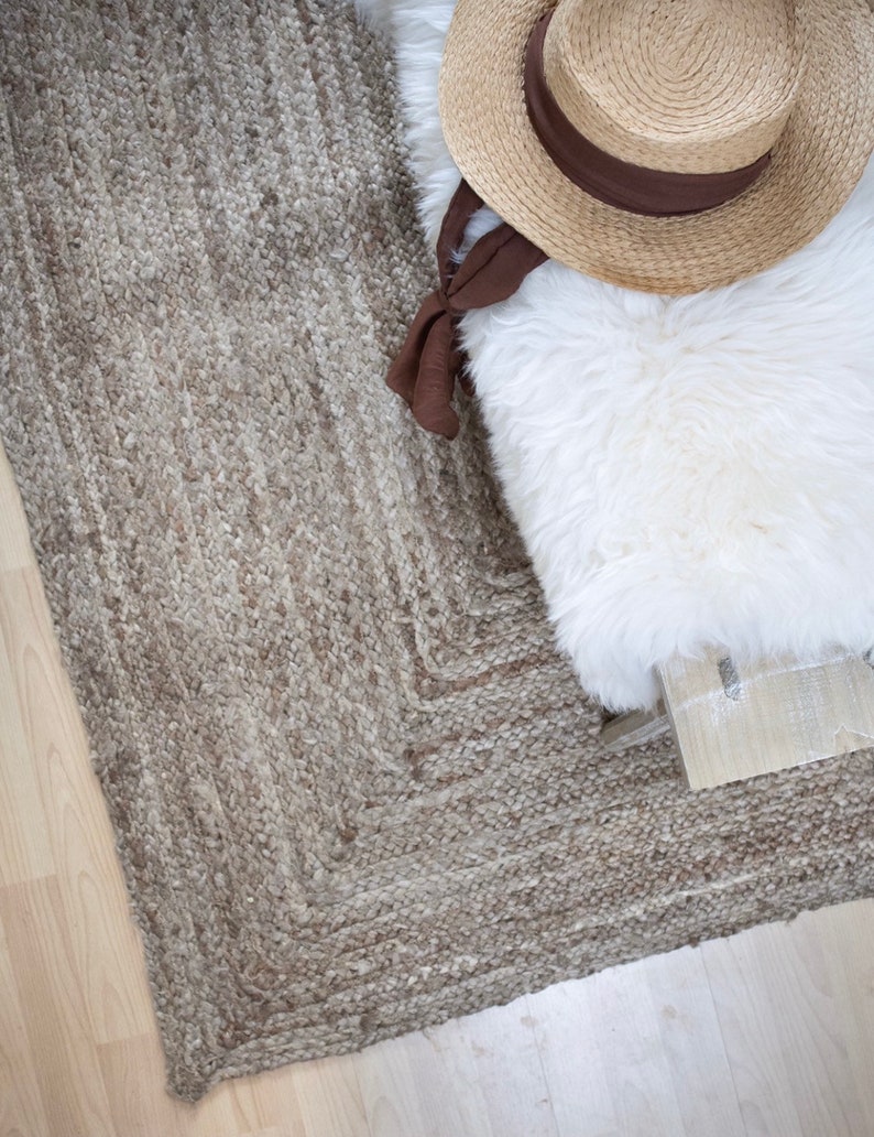 May include: A close-up of a woven jute rug with a light brown color. The rug has a textured surface and a geometric pattern. A white faux fur throw and a straw hat are on top of the rug.