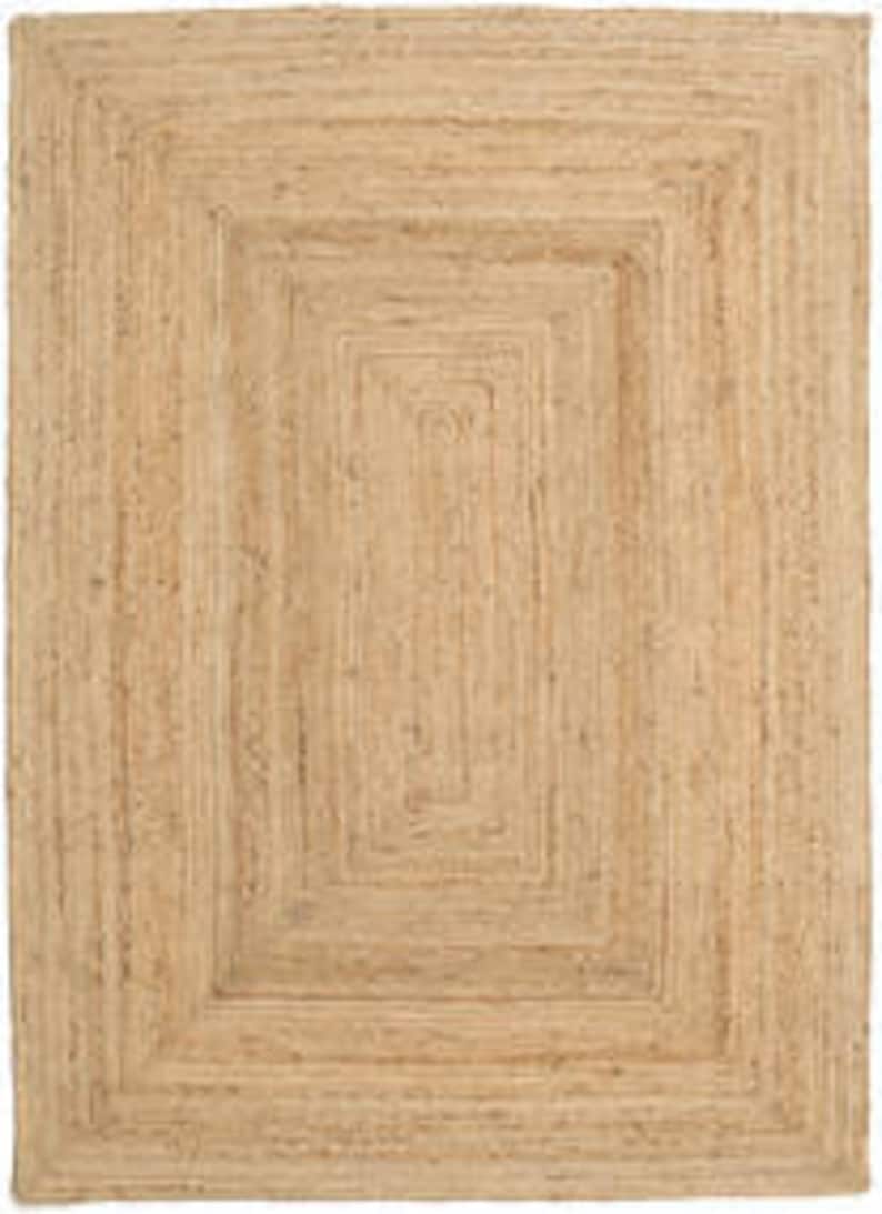 May include: Natural jute rug with a woven design featuring multiple concentric squares. The rug has a light brown color and a rectangular shape.