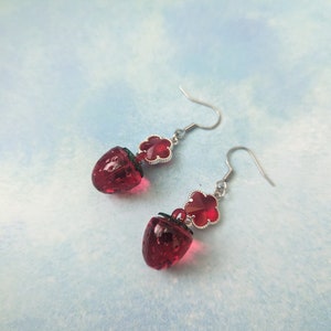 Peut inclure: Une paire de boucles d'oreilles pendantes avec un charme en verre rouge en forme de fraise et un charme en tr&egrave;fle rouge. Les boucles d'oreilles sont argent&eacute;es et ont une fermeture &agrave; crochet.