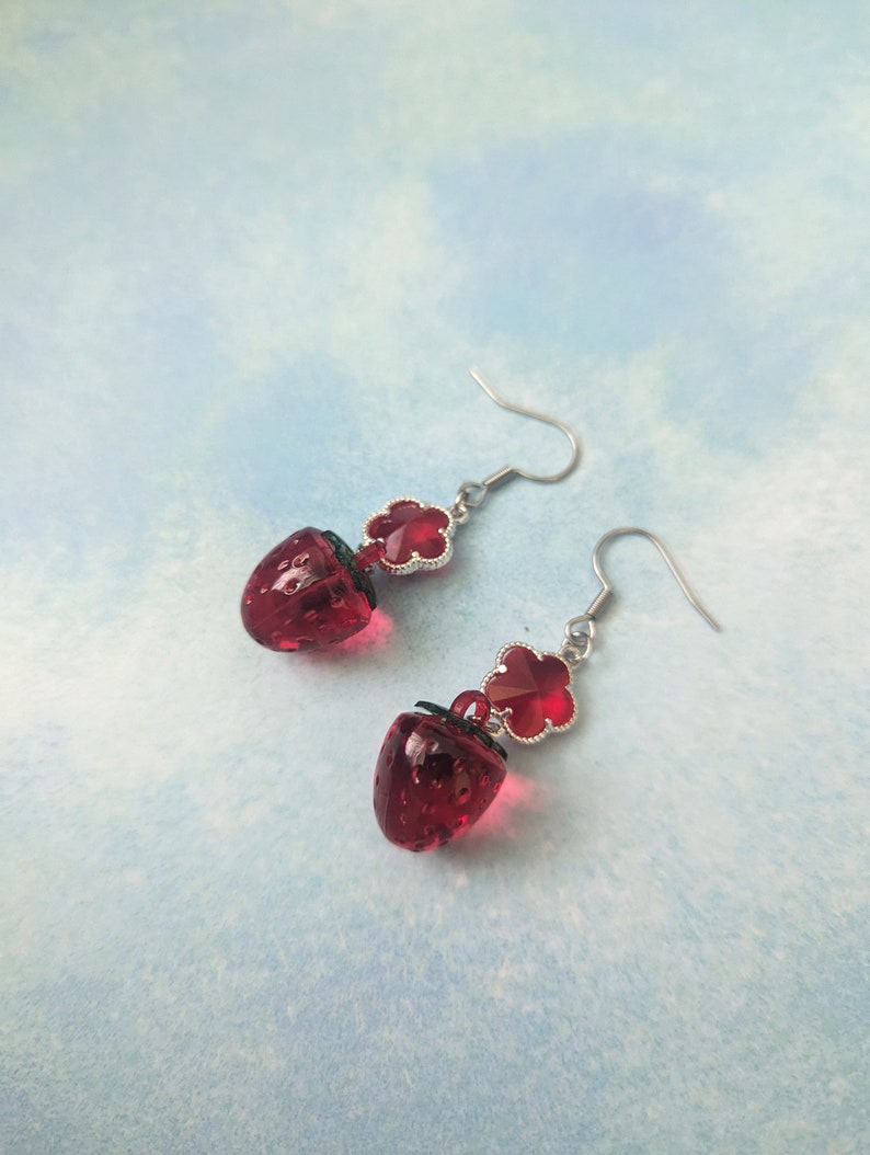 Peut inclure: Une paire de boucles d'oreilles pendantes avec un charme en verre rouge en forme de fraise et un charme en tr&egrave;fle rouge. Les boucles d'oreilles sont argent&eacute;es et ont une fermeture &agrave; crochet.