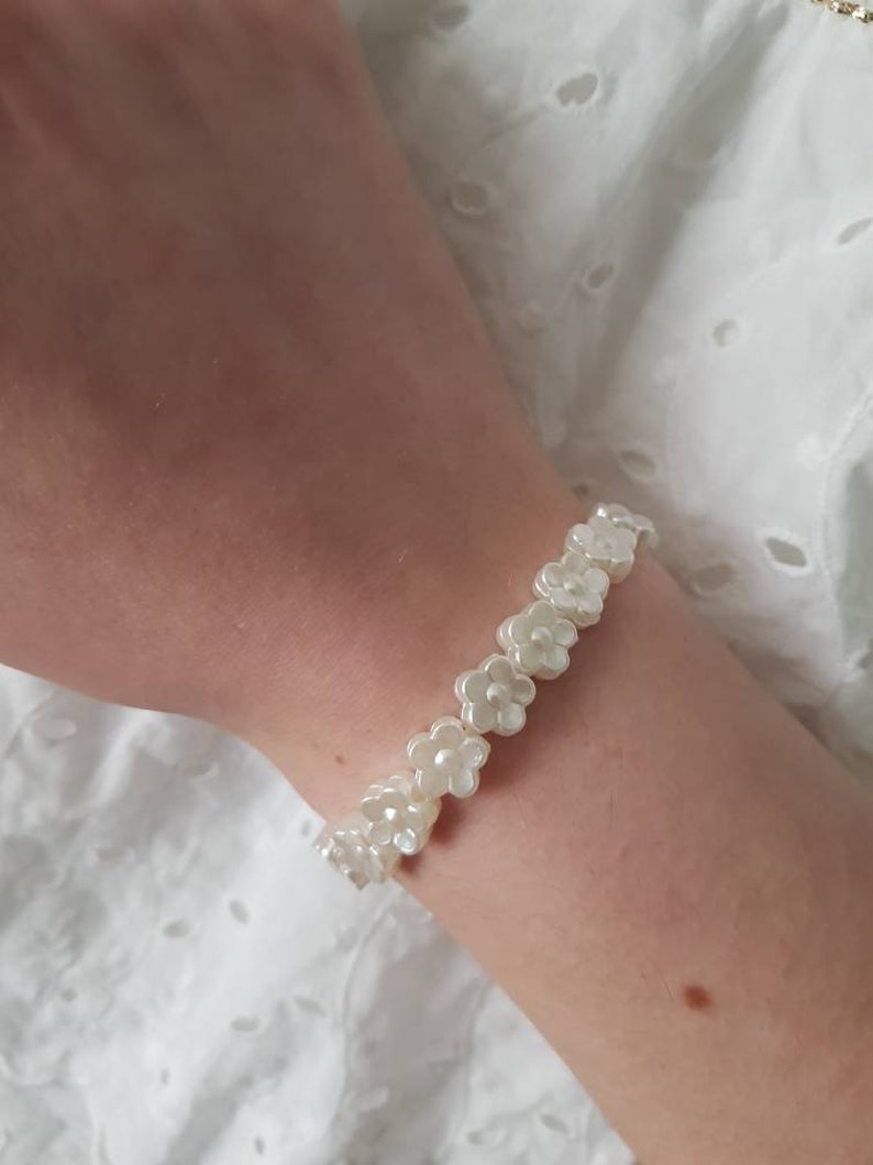 May include: A white bracelet made of small, pearl-like flower charms. The bracelet is on a person's wrist.