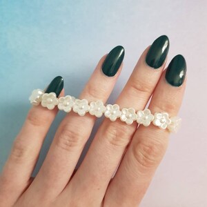 May include: A white pearl flower bracelet with a string of small white flowers. The bracelet is on a hand with dark green nail polish.