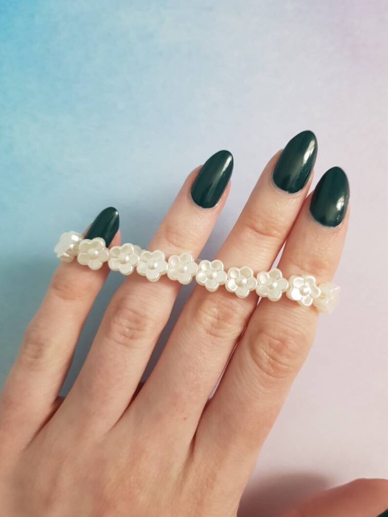 May include: A white pearl flower bracelet with a string of small white flowers. The bracelet is on a hand with dark green nail polish.