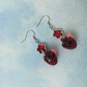 Peut inclure: Une paire de boucles d'oreilles en argent avec des breloques en verre rouge en forme de fraise et des accents de fleurs rouges.