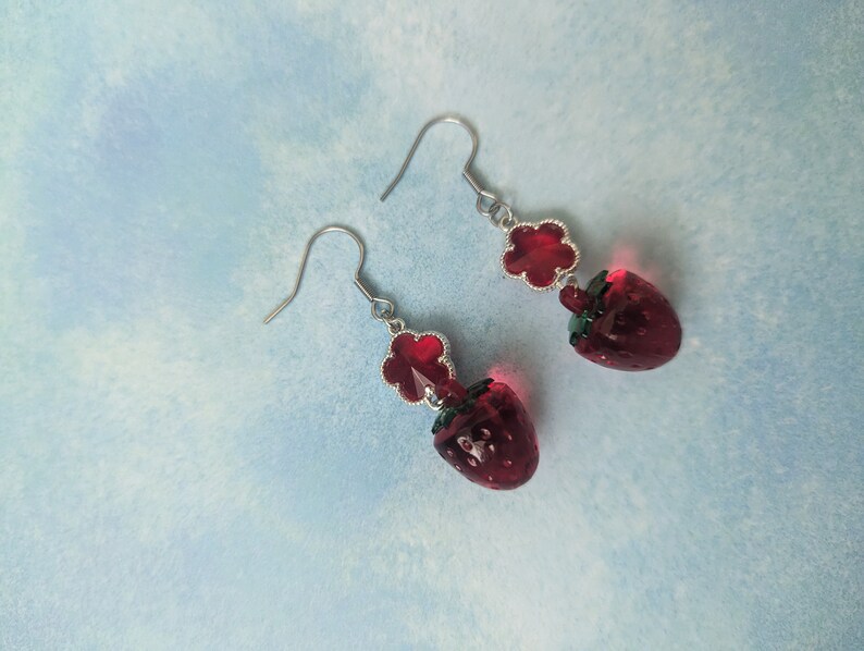 Peut inclure: Une paire de boucles d'oreilles en argent avec des breloques en verre rouge en forme de fraise et des accents de fleurs rouges.