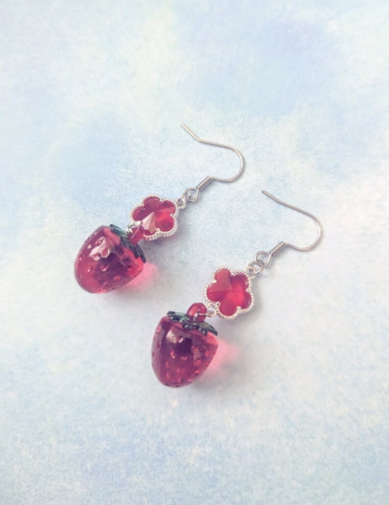 Peut inclure: Une paire de boucles d'oreilles pendantes avec un charme de fraise rouge et un charme de fleur en cristal rouge. Les boucles d'oreilles sont en m&eacute;tal argent&eacute;.