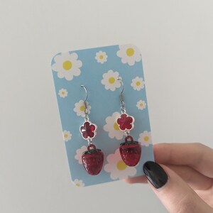 Peut inclure: Une paire de boucles d'oreilles pendantes avec un charme de fleur rouge et un charme de fraise rouge. Les boucles d'oreilles sont sur un fond bleu avec des marguerites blanches.