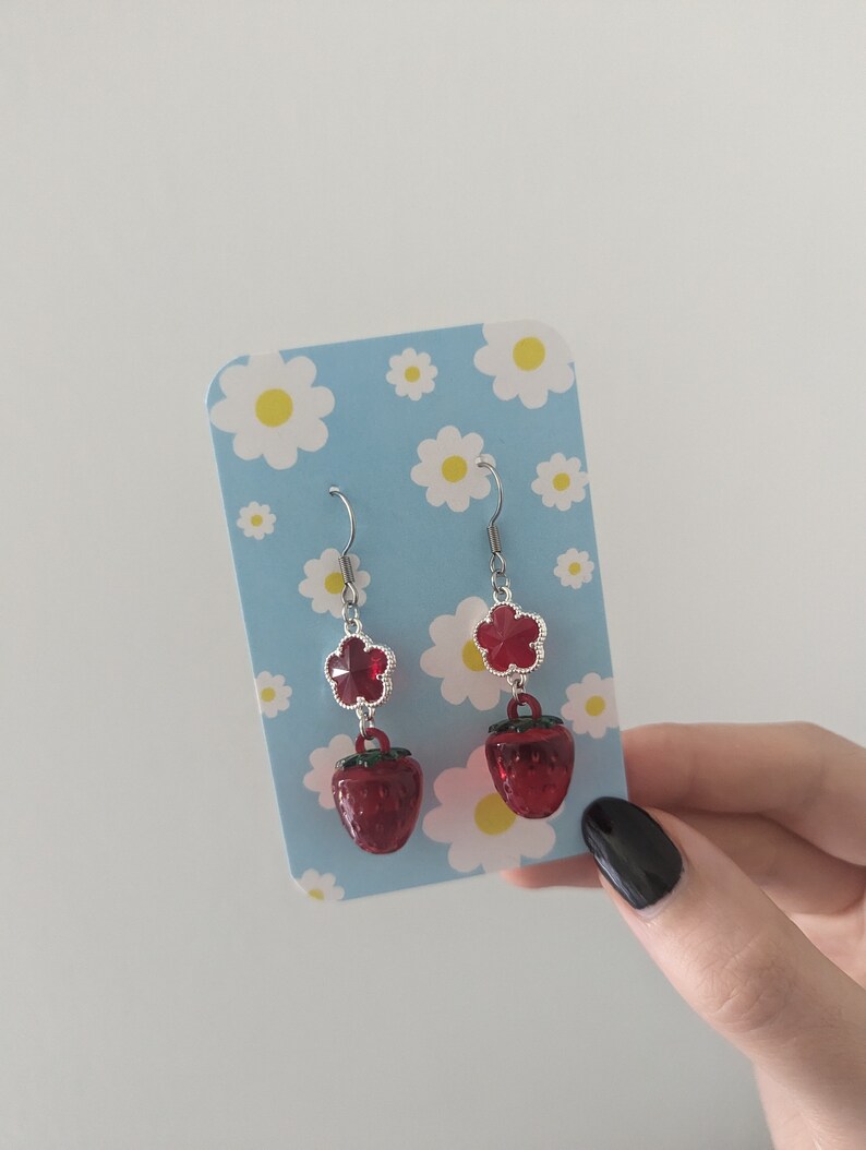Peut inclure: Une paire de boucles d'oreilles pendantes avec un charme de fleur rouge et un charme de fraise rouge. Les boucles d'oreilles sont sur un fond bleu avec des marguerites blanches.