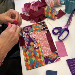 May include: A colourful patchwork quilt being assembled, with patterned fabric squares and burgundy strips. A pair of purple scissors and a blue mug are on a white surface. The quilt features floral and geometric designs. Various fabric pieces are scattered around.