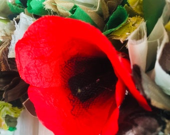 May include: A close-up of a handmade wreath featuring a vibrant red poppy with a black center. The wreath is adorned with various fabric scraps in shades of green, beige, and brown, creating a textured, rustic appearance. The poppy is the focal point.
