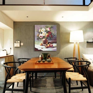 May include: A painting of a vase filled with white and pink roses, with a bowl of fruit on a white tablecloth. The painting is hanging on a wall in a dining room with a wooden table and chairs.