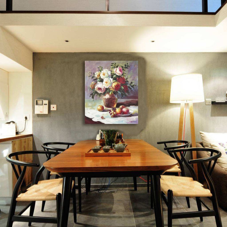 May include: A painting of a vase filled with white and pink roses, with a bowl of fruit on a white tablecloth. The painting is hanging on a wall in a dining room with a wooden table and chairs.