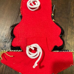 May include: A red felt decorative item with black accents and white pipe cleaner details. Gold and black trim adorns the bottom edge. The item has a unique shape and design, possibly a costume piece or ornament.