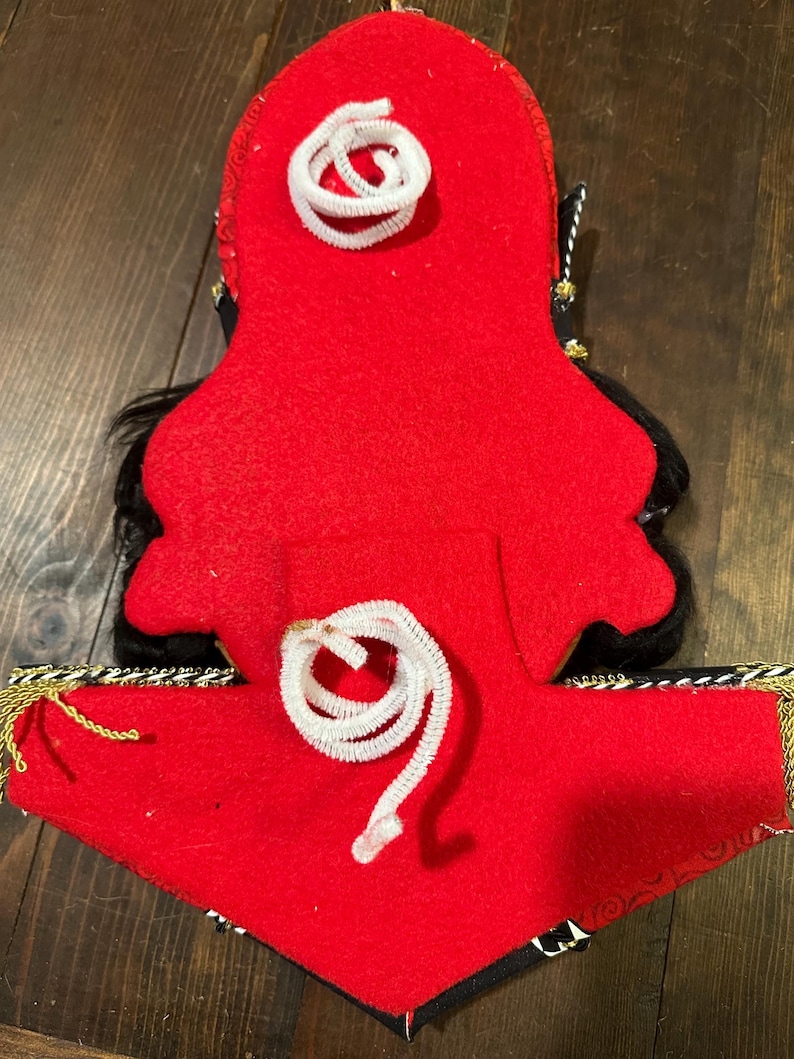 May include: A red felt decorative item with black accents and white pipe cleaner details. Gold and black trim adorns the bottom edge. The item has a unique shape and design, possibly a costume piece or ornament.