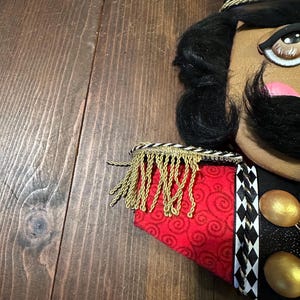 May include: A decorative nutcracker with a black mustache, black hair, and a gold and black hat. The nutcracker has a red and gold patterned jacket with gold fringe and black and white diamond-patterned trim. Two gold orbs are visible.