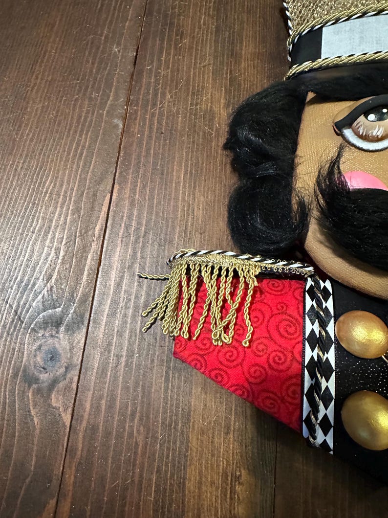 May include: A decorative nutcracker with a black mustache, black hair, and a gold and black hat. The nutcracker has a red and gold patterned jacket with gold fringe and black and white diamond-patterned trim. Two gold orbs are visible.