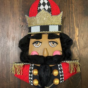 May include: A decorative nutcracker face with a red and black hat, gold glitter crown, and black hair and beard. The face has painted features, including pink cheeks and a brown nose. Gold trim and accents complete the design.