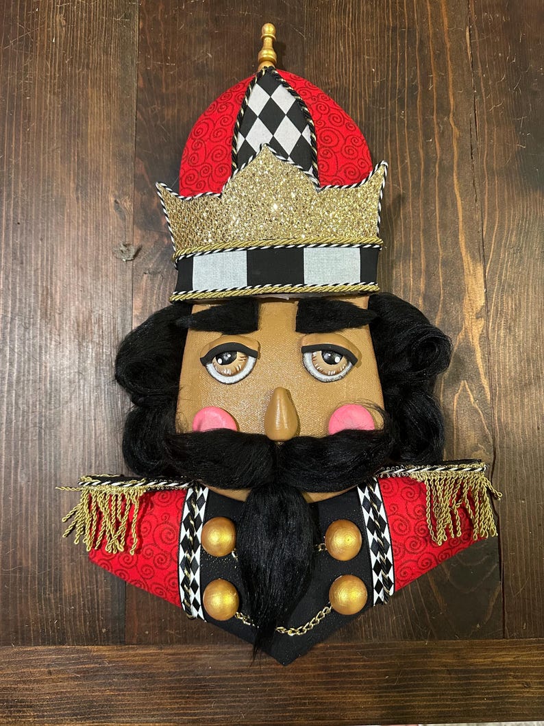 May include: A decorative nutcracker face with a red and black hat, gold glitter crown, and black hair and beard. The face has painted features, including pink cheeks and a brown nose. Gold trim and accents complete the design.