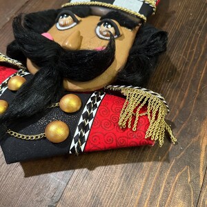 May include: A decorative nutcracker head with a black mustache and hair, wearing a gold crown and a black, red, and white uniform. The uniform has gold accents and fringe.