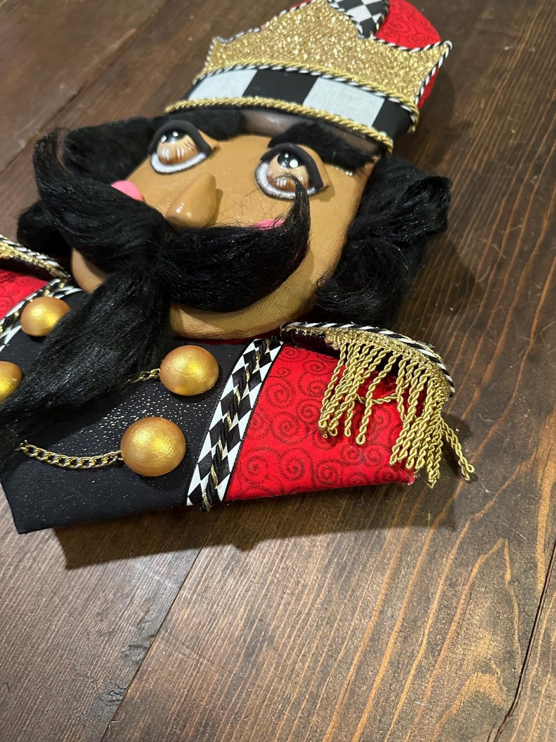 May include: A decorative nutcracker head with a black mustache and hair, wearing a gold crown and a black, red, and white uniform. The uniform has gold accents and fringe.