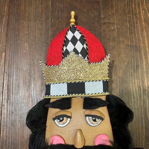 May include: A decorative nutcracker with a tan face, black hair, and a black mustache. The nutcracker wears a red and black hat with a gold crown. The crown is trimmed with gold and black accents. The nutcracker has pink cheeks and a long nose.