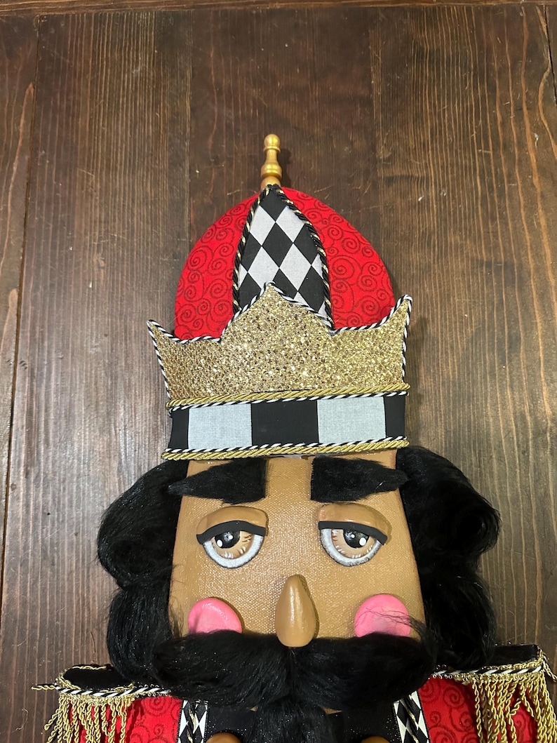 May include: A decorative nutcracker with a tan face, black hair, and a black mustache. The nutcracker wears a red and black hat with a gold crown. The crown is trimmed with gold and black accents. The nutcracker has pink cheeks and a long nose.