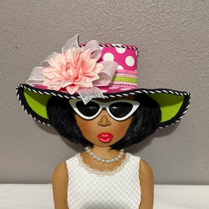 May include: A decorative figure of a woman wearing a pink polka dot hat with a large pink flower and white sunglasses. The figure has a white dress and a pearl necklace. The hat has a green brim and a black and white trim.