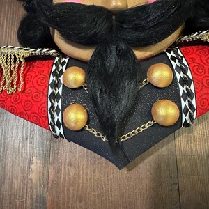 May include: Close-up of a decorative nutcracker head with a dark complexion, black mustache, and black hair. The nutcracker wears a red jacket with gold trim and black accents. Gold spheres are connected by a gold chain.