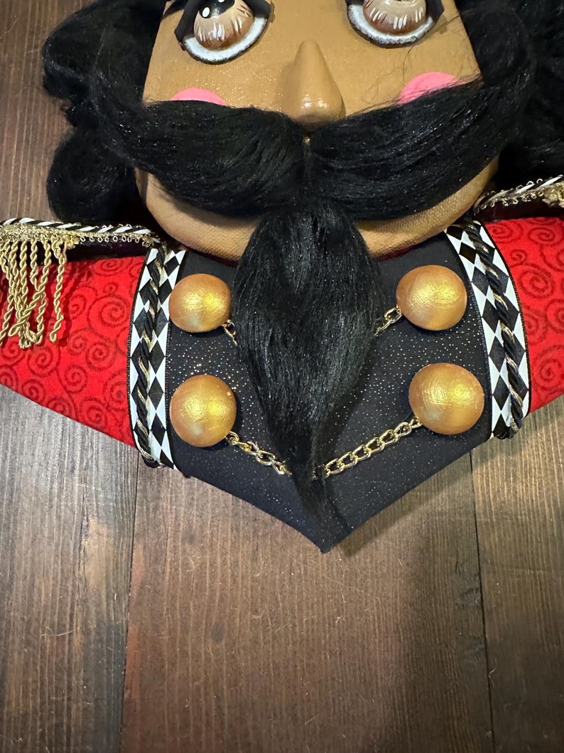 May include: Close-up of a decorative nutcracker head with a dark complexion, black mustache, and black hair. The nutcracker wears a red jacket with gold trim and black accents. Gold spheres are connected by a gold chain.
