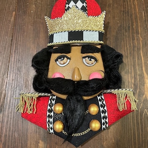 Puede incluir: Una cara decorativa de cascanueces con un sombrero rojo y negro, una corona dorada y cabello y bigote negros. El cascanueces tiene una cara bronceada con mejillas rosadas y un uniforme negro con botones y adornos dorados.