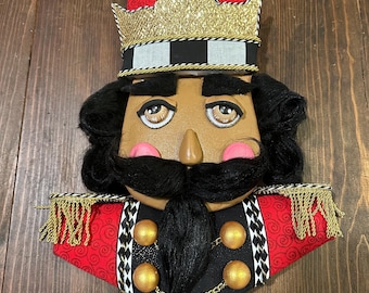 May include: A decorative nutcracker face with a red and black hat, gold crown, and black hair and mustache. The nutcracker has a tan face with pink cheeks and a black uniform with gold buttons and trim.