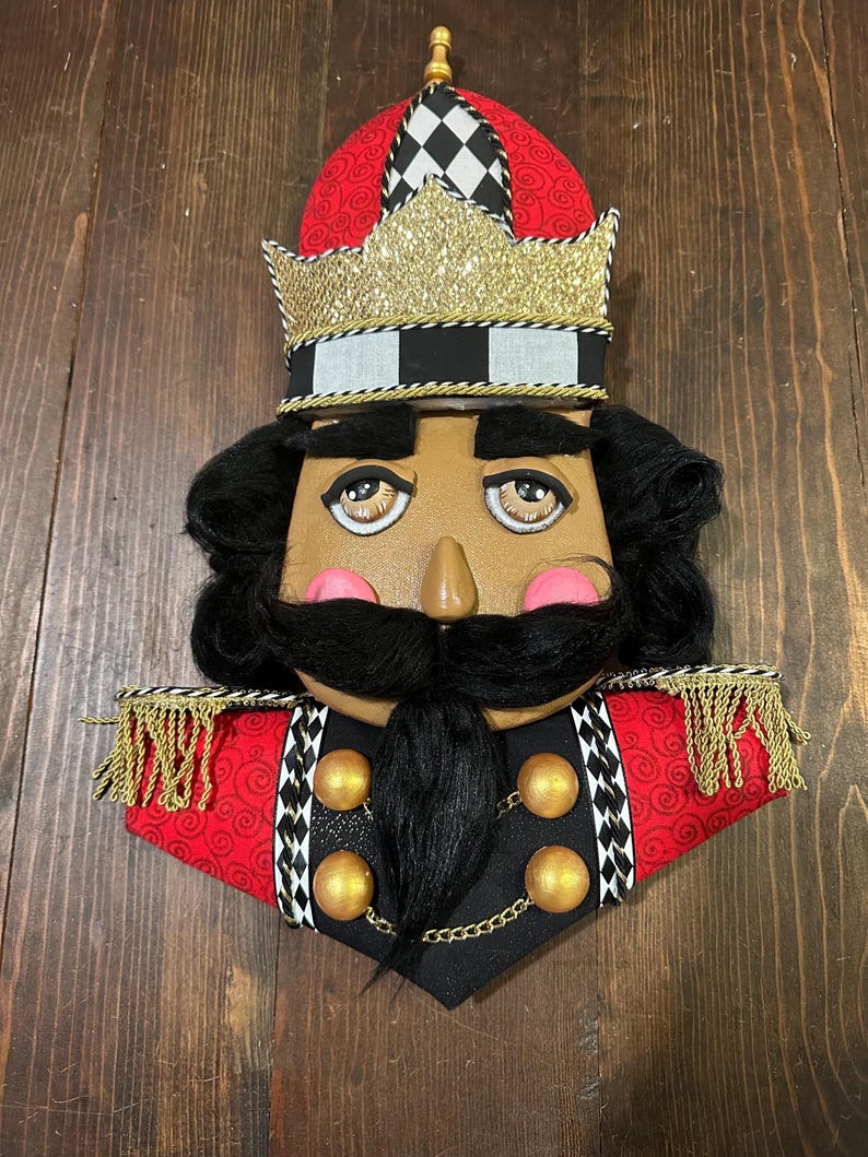 May include: A decorative nutcracker face with a red and black hat, gold crown, and black hair and mustache. The nutcracker has a tan face with pink cheeks and a black uniform with gold buttons and trim.