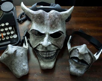 May include: A collection of three gray, textured masks with black eye holes and straps. The central mask has large horns and a wide, toothy grin. The other two masks have different facial expressions. A vintage typewriter is in the background.