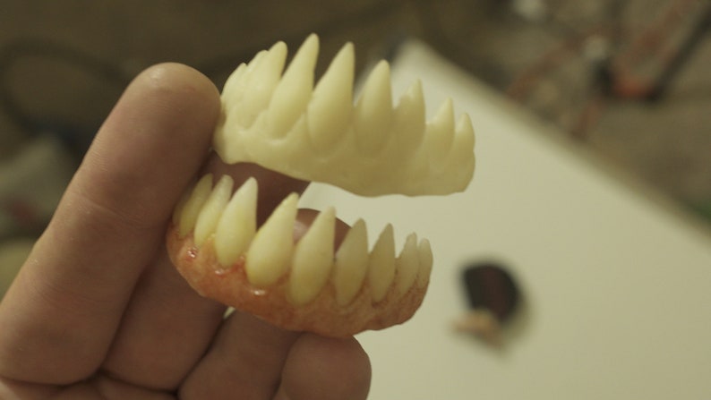 Pu&ograve; includere: Un paio di denti da vampiro in plastica bianca con una base rossa. I denti sono affilati e appuntiti e sono tenuti nella mano di una persona.