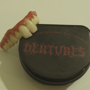 Pu&ograve; includere: Un set di denti finti di colore rosa e bianco. I denti sono in una custodia nera con la parola "DENTURES" stampata in rosa.