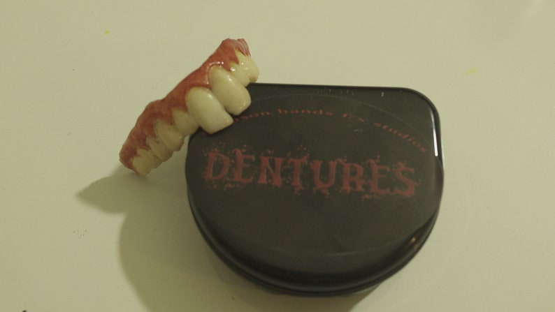 Pu&ograve; includere: Un set di denti finti di colore rosa e bianco. I denti sono in una custodia nera con la parola "DENTURES" stampata in rosa.