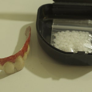 May include: A set of fake teeth in a plastic case with a small bag of adhesive. The teeth are made of a white plastic and have a pink gum line. The adhesive is in a clear plastic bag.